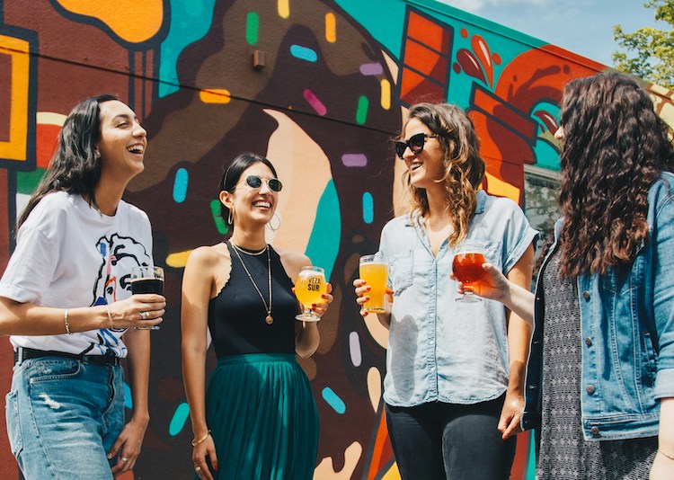 Four women stand in front of brightly painted wall drinking
